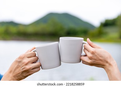 Asian Couple hand toasting coffee cup while having breakfast and drinking coffee together in the morning. Man and woman enjoy outdoor lifestyle camping in forest mountain on summer travel vacation - Powered by Shutterstock