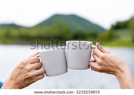 Asian Couple hand toasting coffee cup while having breakfast and drinking coffee together in the morning. Man and woman enjoy outdoor lifestyle camping in forest mountain on summer travel vacation