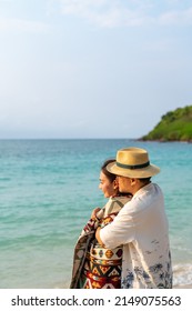 Asian Couple Husband Cover His Wife With A Blanket And Hugging Each Other While Walking On Tropical Beach Together At Summer Sunset. Happy Family Relax And Enjoy Outdoor Lifestyle On Travel Vacation.