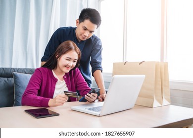 Asian Couple Online Shopping Ecommerce Together From Home Modern Living Room Using Phone Computer Laptop Tablet Payment Paying Transaction Money Transfer Credit Debit Card Smiling Happy Joyfully 