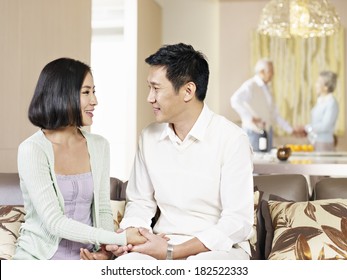 Asian Couple Sitting On Couch And Looking At Each Other With Their Senior Parents In The Background