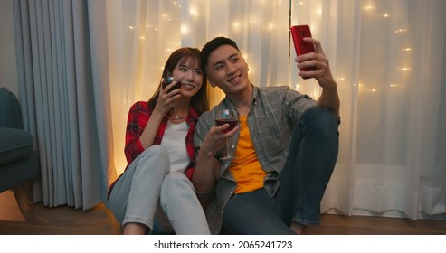 Asian Couple Sitting On Floor Take Selfie With Red Wine While Celebrating Anniversary At Night In Living Room