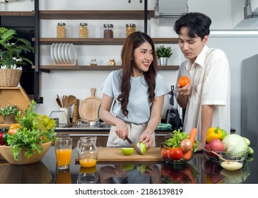 Asian Couple Spend Time Together In The Kitchen. Young Woman Cutting Green Apple On A Wooden Chop Board While Her Boyfriend Stand Beside Her With An Orange In His Hand. Fruit Juice Are On The Counter.