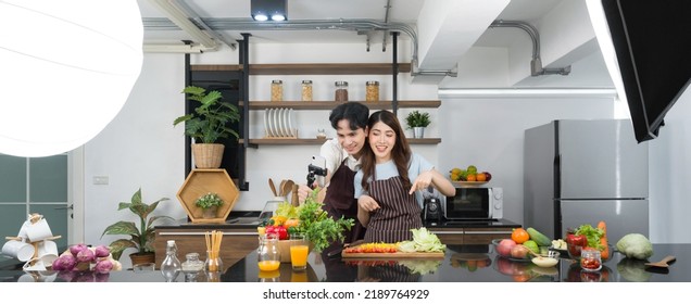 Asian Couple Spend Time Together In The Kitchen. Young Woman In Apron Cooking Salad Dish While His Boyfriend Recording Vlog Video For Social Blogger. Photography Studio Lights Set Up In The Kitchen.