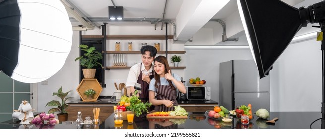 Asian Couple Spend Time Together In The Kitchen. Young Woman In Apron Cooking Salad Dish While His Boyfriend Recording Vlog Video For Social Blogger. Photography Studio Lights Set Up In The Kitchen.