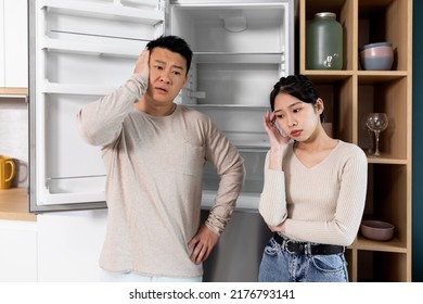 Asian Couple Standing Next To Open Empty Fridge In Kitchen At Home, Touching Their Faces. Poor Chinese Family Have Nothing To Eat, Copy Space. Financial Hangover While Crisis, Poverty Concept