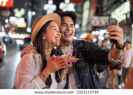 Asian couple tourist backpacker doing selfie and recording a video blog while eating food from street stall in night market with crowd of people at Yaowarat road, Bangkok