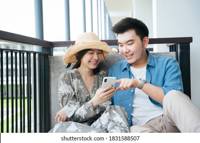 Asian Couple Traveler Sitting At Hotel Balcony And Using Smartphone.