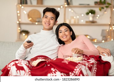 Asian Couple Watching Christmas Movies On TV Eating Popcorn Sitting On Sofa At Home, Covered With Blanket. Family Enjoying Xmas Eve And Holidays Television Show In Cozy Living Room Concept