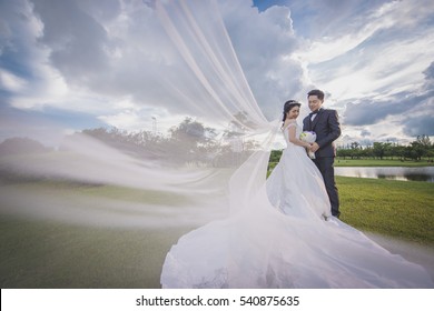 Asian Couple Wedding Outdoor Style Under A Cloudy Sky On A Green Grass Field With Bride's Suit's Part Spreading In The Air