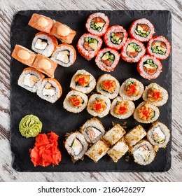 Asian Cuisine. Large Sushi Set Of Various Sushi Rolls Is Served On Black Square Metal Slate. Top View Of Different Rolls With Variety Of Fresh Ingredients And Wasabi With Ginger On Edge.