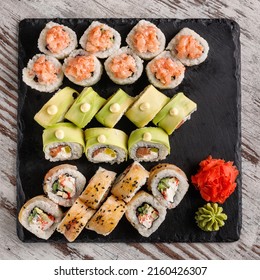 Asian Cuisine. Large Sushi Set Of Various Sushi Rolls Is Served On Black Square Metal Slate. Top View Of Different Rolls With Variety Of Fresh Ingredients And Wasabi With Ginger On Edge.