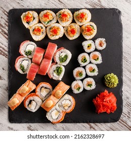 Asian Cuisine. Large Sushi Set Of Various Sushi Rolls Is Served On Black Square Metal Slate. Top View Of Different Rolls With Variety Of Fresh Ingredients And Wasabi With Ginger On Edge.