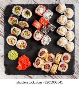 Asian Cuisine. Large Sushi Set Of Various Sushi Rolls Is Served On Black Square Metal Slate. Top View Of Different Rolls With Variety Of Fresh Ingredients And Wasabi With Ginger On Edge.