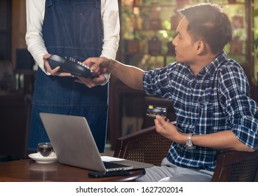 Asian Customer Using Credit Card With EMV Chip Technology To Pay A Waitress For Coffee Purchase At Table In Cafe.