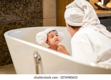 Asian Cute Baby In A Bathrobe And Hair Wrapped In Towel Pointing On Chin And Having Fun With Her Mother In A Bathtub