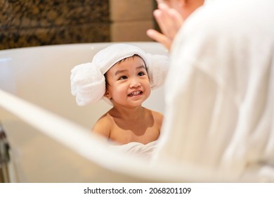 Asian Cute Baby Girl In A Bathrobe And Hair Wrapped In Towel Having Fun With Her Mother In A Bathtub