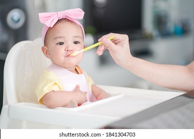 Asian Cute Baby Girl In A Pink Bib And Hair Bow Sitting On A White High Chair. Her Mother Giving Healthy Food By Spoon In The Morning At Home