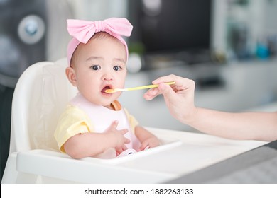 Asian Cute Baby Girl In A Pink Bib And Hair Bow Sitting On A White High Chair. Her Mother Giving Healthy Food By Spoon In The Morning At Home