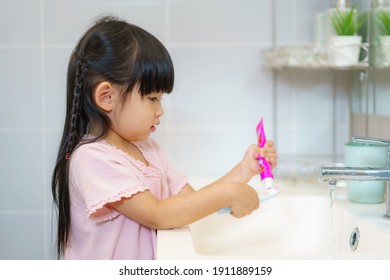 Asian Cute Child Girl Or Kid Squeeze The Toothpaste Into Toothbrush Before Brushing Her Teeth In The Bathroom. Dental Hygiene Healthcare Concept.