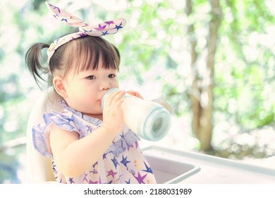 Asian Cute Happy Baby Drinking Her Milk From Bottle On A Chair. 1 Year 6 Months Baby Handing Flower Use As Concept Of Mood, Summer, Health, Baby Development Or Kid Department In Hospital.