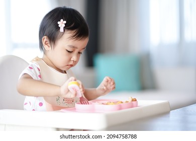 Asian cute little girl holding fork and trying to eat food sitting on the white high chair in the morning at home with copy space.