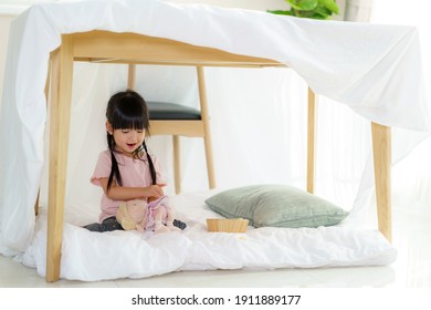 Asian cute little girl play her doll while sitting in a blanket fort in living room at home for perfect hideout away from their other family members and for them to play imaginatively.