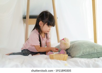 Asian Cute Little Girl Play Her Doll And Feeding Snack While Sitting In A Blanket Fort In Living Room At Home For Perfect Hideout Away From Their Other Family Members