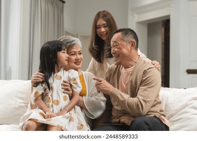 Asian cute little girl sitting on sofa with grandparent and young mother, talking, smiling together. Happy grandchild listen to grandfather fun story at home. Multi generation family relaxing together - Powered by Shutterstock