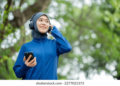 Asian cute muslim woman wearing sportswear and a blue hijab Working out and wearing earphones listening to music on her phone outdoors at the park in the morning with a fresh feeling. - Powered by Shutterstock