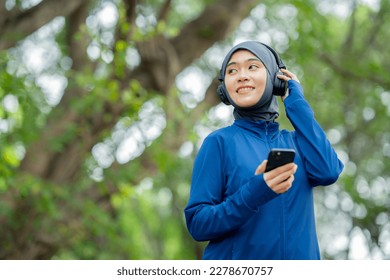 Asian cute muslim woman wearing sportswear and a blue hijab Working out and wearing earphones listening to music on her phone outdoors at the park in the morning with a fresh feeling. - Powered by Shutterstock