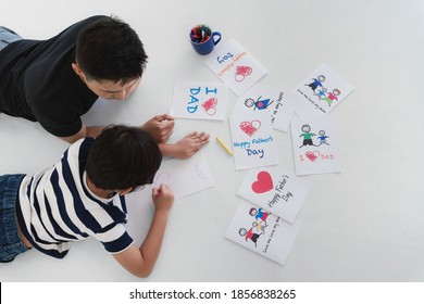 Asian Dad And Son Lying On Floor Together Drawing Gift Card For Father Day At Home. Concept Of Dad And Son Relationship And Father Day