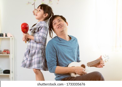 Asian Daddy And Cute Little Girl Is Singing And Play The Ukulele And Maraca Together In The Room With Happy Moment, Concept Of Life At Home And Active Learning Activity For Kid And Role Of Parent.