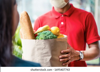 Asian deliver man wearing face mask in red uniform handling bag of food, fruit, vegetable give to female costumer in front of the house. Postman and express grocery delivery service during covid19.