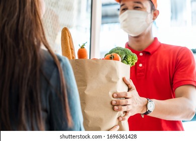 Asian deliver man wearing face mask in red uniform handling bag of food, fruit, vegetable give to female costumer in front of the house. Postman and express grocery delivery service during covid19. - Shutterstock ID 1696361428
