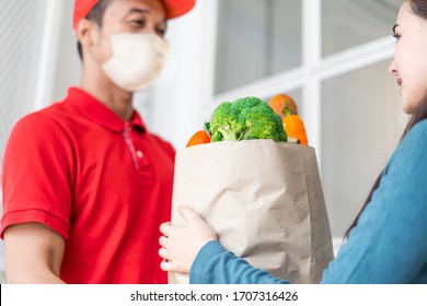 Asian Deliver Man Wearing Face Mask In Red Uniform Handling Bag Of Food, Fruit, Vegetable Give To Female Costumer In Front Of The House. Postman And Express Grocery Delivery Service During Covid19.