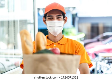 Asian Deliver Man Wearing Face Mask In Orange Uniform Holding Bag Of Food, Groceries, Fruit Standing In Front Of Customer Home. Postman And Express Grocery Delivery Service During Covid19 Pandemic.