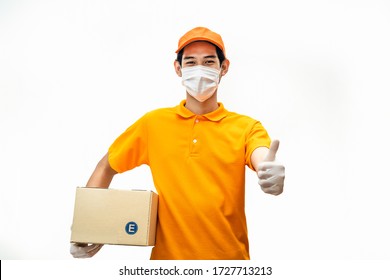 Asian deliver man wearing face mask in orange uniform holding box of food, groceries gives thumbs up standing in white isolated background. Postman and express delivery service during covid19 pandemic
