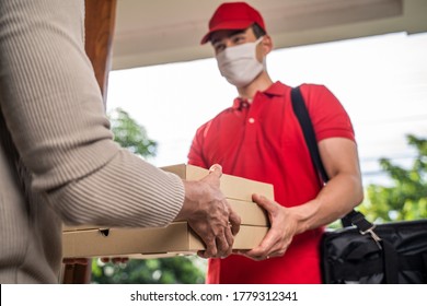 Asian deliver man wearing protective face mask in red uniform handling box of food, pizza give to female costumer in front of the house. Postman and express grocery delivery service during covid19.
