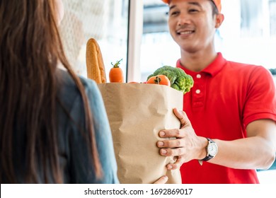 Asian deliver man worker in red color uniform handling bag of food, fruit, vegetable give to young beautiful female costumer in front of the house. Postman and express grocery delivery service concept - Shutterstock ID 1692869017