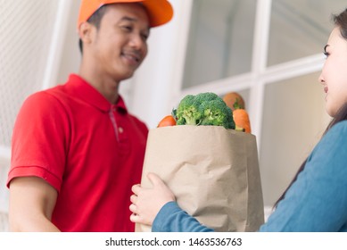 Asian delivery man in red color uniform handling bag of foods or vegetables to young beautiful female costumer in front of the house. Postman or express delivery service concept. Focus on girl eyes. - Shutterstock ID 1463536763