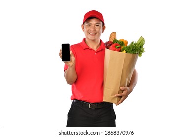 Asian Delivery Man In Red Uniform Carry Grocery Bag And Showing Mobile Phone Isolated On White Background       