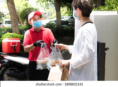 Asian Delivery Man Wearing Face Mask And Gloves In Red Uniform Delivering Bag Of Food And Drink To Recipient During COVID-19 Outbreak