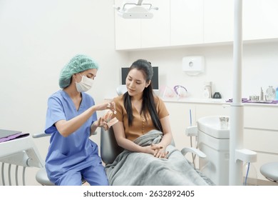 Asistente dental asiático con máscara quirúrgica y uniforme usando el modelo dental para explicar los pasos del tratamiento a la paciente femenina sentada en una silla dental durante la consulta en la sala de exámenes moderna limpia.