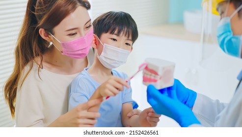 Asian Dentist With Face Shield And Mask Teach Boy How Use Toothbrush Brushing Teeth Use Denture Model When Mother Sit By In Dental Clinic - Kid Practice It