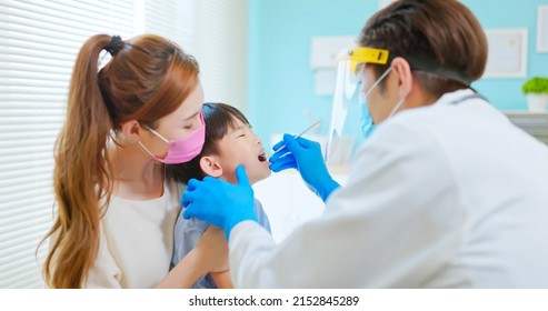 Asian Dentist With Face Shield And Mask Use Mouth Mirror To Exam Toothache Boy When Mother Sit By In Dental Clinic - He Looked Scared