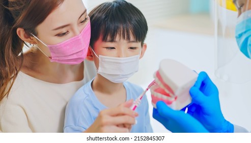 Asian Dentist With Face Shield And Mask Teach Boy How To Toothbrush Brushing Teeth Use Denture Model When Mother Sit By In Dental Clinic - Kid Practice It