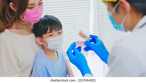 Asian Dentist With Face Shield And Mask Teach Boy Knowledge About Braces Use Denture Model When Mother Sit By In Dental Clinic