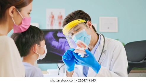 Asian Dentist With Face Shield And Mask Teach Boy Knowledge About Braces Use Denture Model When Mother Sit By In Dental Clinic