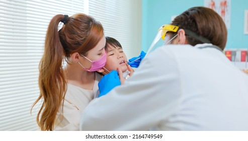 Asian Dentist With Face Shield And Mask Use Mouth Mirror To Exam Toothache Boy When Mother Sit By In Dental Clinic - He Looked Scared